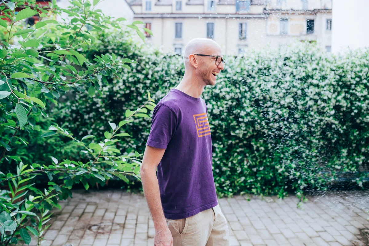 A smiling man in a purple Sobrynth tee walking past a green hedge